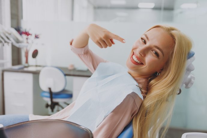 Woman smiling and pointing to her healthy teeth while relaxing in a dental chair at Integrated Aesthetic Dentistry in New York City